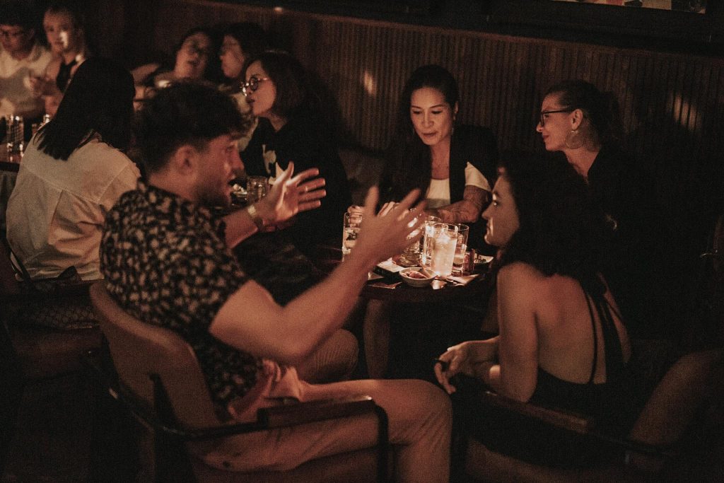 426621221 877805467687050 952382951408965682 n A dimly lit bar scene featuring patrons seated at tables, engaged in conversation and enjoying their drinks.