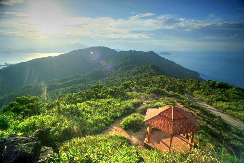 The photo captures a breathtaking view from Ban Co Peak, with the sun casting a golden light over the vast landscape. From this vantage point, the city of Da Nang stretches out below, framed by the sparkling blue sea and lush green hills. The warm sunlight enhances the natural beauty, creating a peaceful and awe-inspiring scene that leaves visitors in awe of the stunning panorama.
