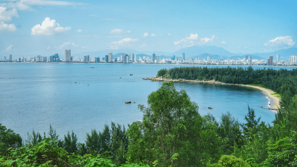 The photo captures a breathtaking view from Ban Co Peak, with the sun casting a golden light over the vast landscape. From this vantage point, the city of Da Nang stretches out below, framed by the sparkling blue sea and lush green hills. The warm sunlight enhances the natural beauty, creating a peaceful and awe-inspiring scene that leaves visitors in awe of the stunning panorama.