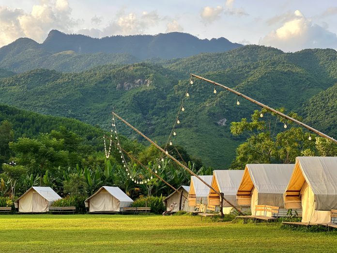 A tranquil view of Yen Retreat featuring lush green fields, the gentle Cu De River, and majestic mountains under a clear blue sky