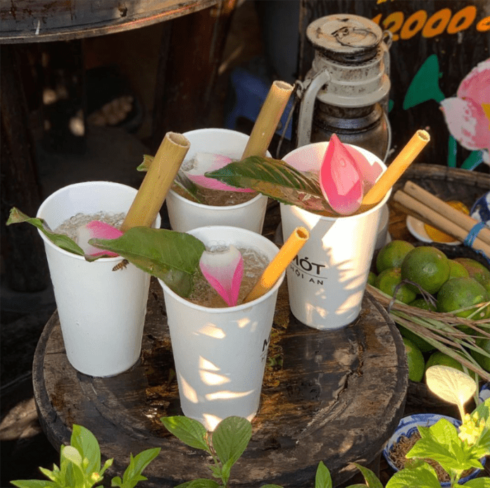 Four cups of Mót herbal tea in paper cups, each adorned with a lotus petal, placed on a tray and photographed in front of the shop in Hoi An Ancient Town. This image captures the cultural beauty and charm of Hoi An's famous drink, perfect for highlighting local flavors and traditions