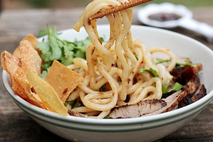 A pair of chopsticks lifts the thick, chewy noodles from a bowl of Cao Lầu, creating an enticing and visually appealing scene. This image highlights the dish's texture and inviting colors, evoking a sense of craving and showcasing the deliciousness of this iconic Hoi An specialty.