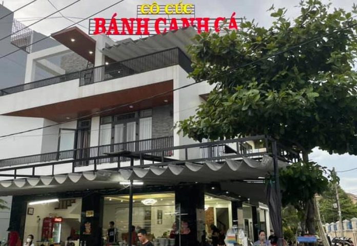Hình ảnh mô tả quán bánh canh cô Cúc từ phía trước.Có cây có mái che có bản hiệu trên cao với chữ Bánh Canh Cá màu đỏ và chữ Cô Cúc màu Vàng