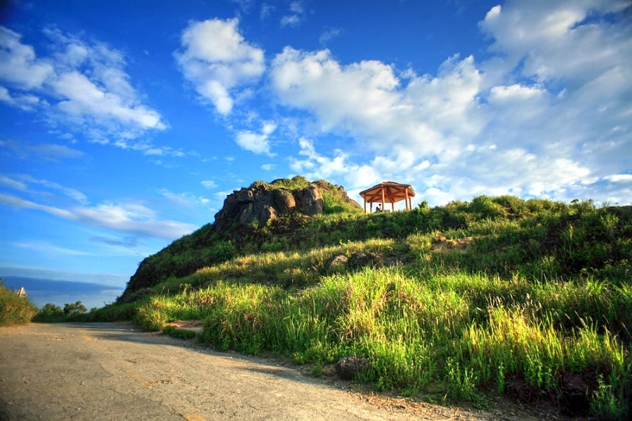 The photo captures a breathtaking view from Ban Co Peak, with the sun casting a golden light over the vast landscape. From this vantage point, the city of Da Nang stretches out below, framed by the sparkling blue sea and lush green hills. The warm sunlight enhances the natural beauty, creating a peaceful and awe-inspiring scene that leaves visitors in awe of the stunning panorama.