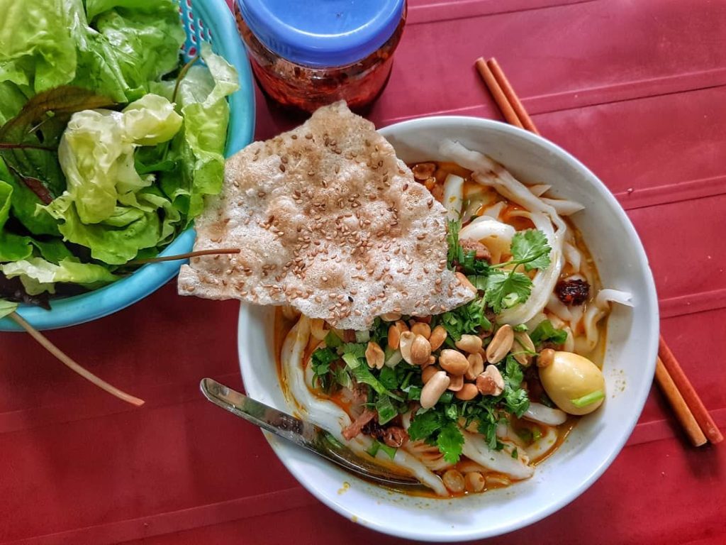 a traditional Vietnamese noodle dish from Hoi An, featuring thick, chewy rice noodles served with tender slices of marinated pork, fresh herbs, and crispy rice crackers. The dish is garnished with bean sprouts and lime, offering a delightful balance of savory, fresh, and slightly tangy flavors, making it a must-try for any food lover