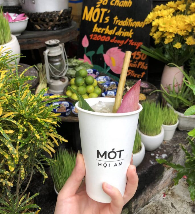 A cup of Mot herbal tea in a paper cup, adorned with a lotus petal, photographed in front of the shop in Hoi An Ancient Town. This iconic beverage and setting capture the charm and cultural essence of Hoi An, perfect for showcasing local flavors and heritage
